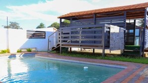A swimming pool with a brick border and a wooden deck with a metal railing. - Phakela Cottages (Jwaneng)