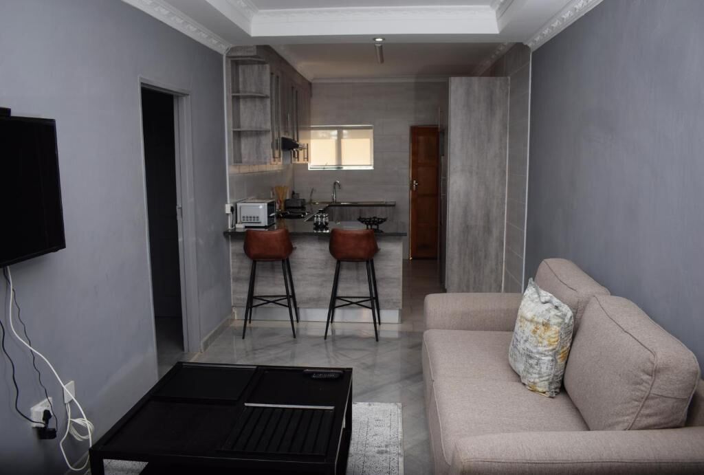 A modern living room with a sofa and a coffee table, a kitchen area with a window, and a TV mounted on the wall.