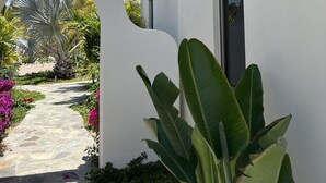 A white building with a stone pathway leading to an archway, surrounded by lush greenery and flowering plants.