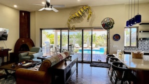 A spacious living area with a fireplace, a ceiling fan, a poolside seating area, and a kitchen with a tiled backsplash.