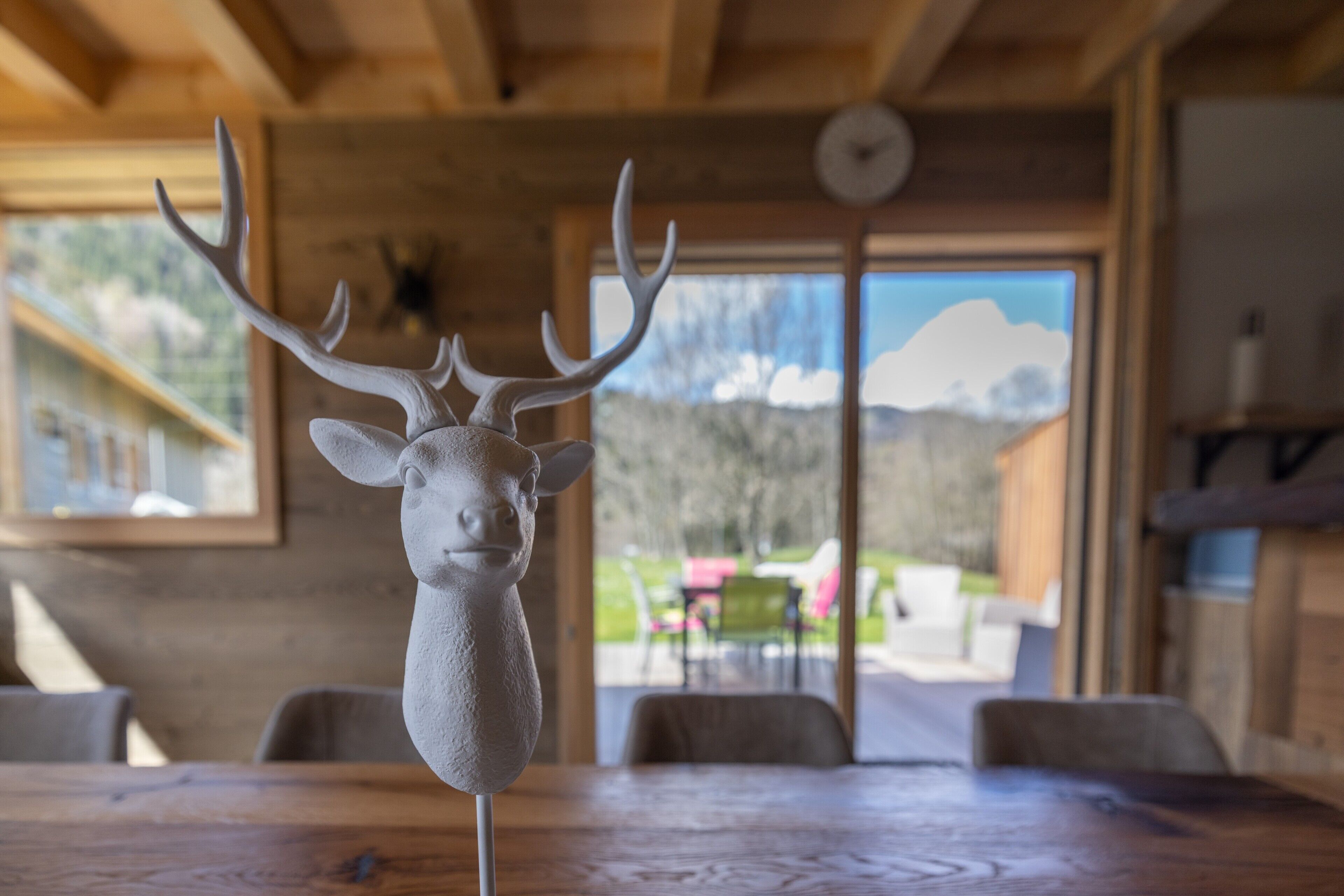 Un interior de madera con una escultura de cabeza de ciervo sobre una mesa, un reloj en la pared y vistas de un paisaje montañoso a través de una ventana.