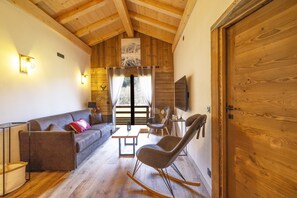 A cozy living room with a wooden ceiling, a sofa, a rocking chair, a television, and a large wooden door.
