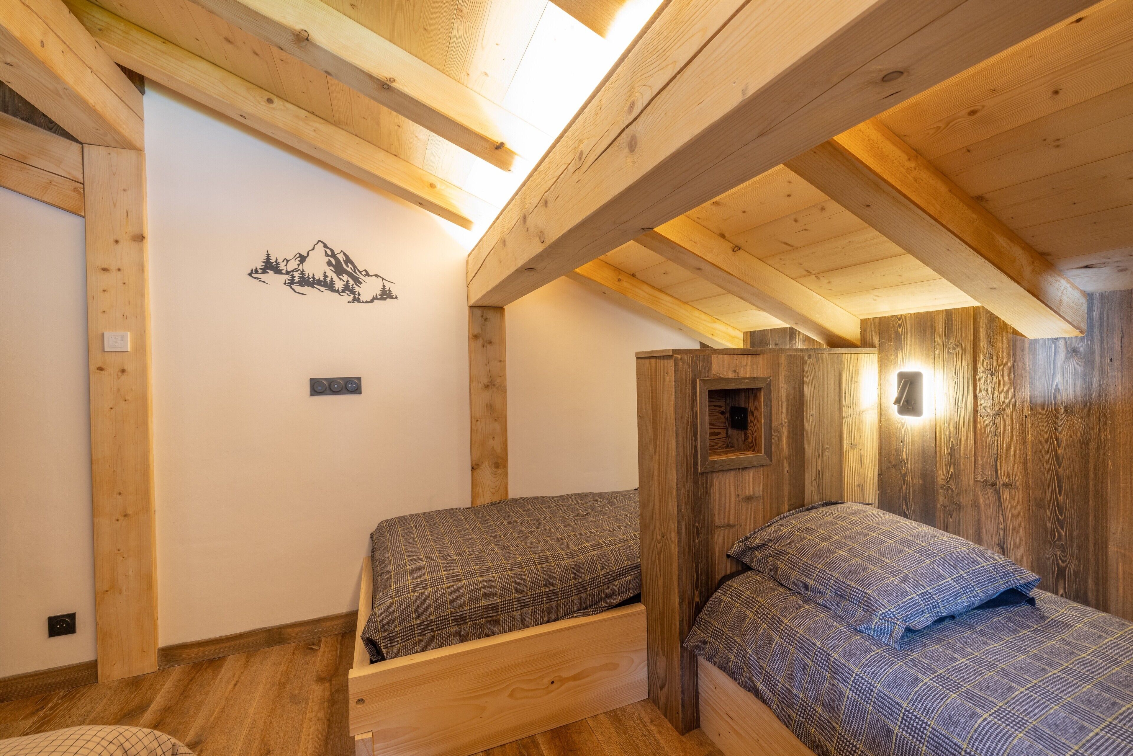 Une pièce avec des poutres apparentes, un lit recouvert d’une courtepointe à carreaux, un lampadaire fixé au mur et une fresque représentant une montagne.