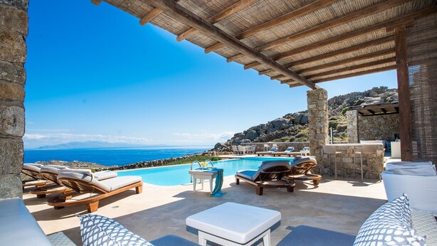 Uma área à beira da piscina com espreguiçadeiras, uma mesa e vista para o mar.