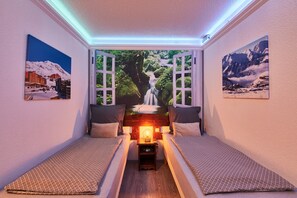 A cozy room with two beds, a nightstand with a lamp, and a view of a waterfall through open windows.