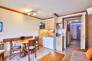 Cocina compacta con microondas, pileta y armarios de madera. Cuenta con un comedor con mesa y sillas, y un sofá con un cojín naranja.