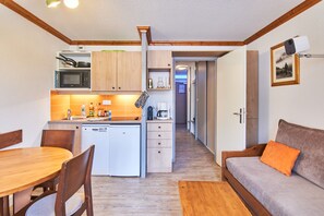 Una cocina compacta con microondas, horno y pileta, una zona de comedor con mesa y sillas, y un espacio de estar con sofá e imagen enmarcada en la pared.