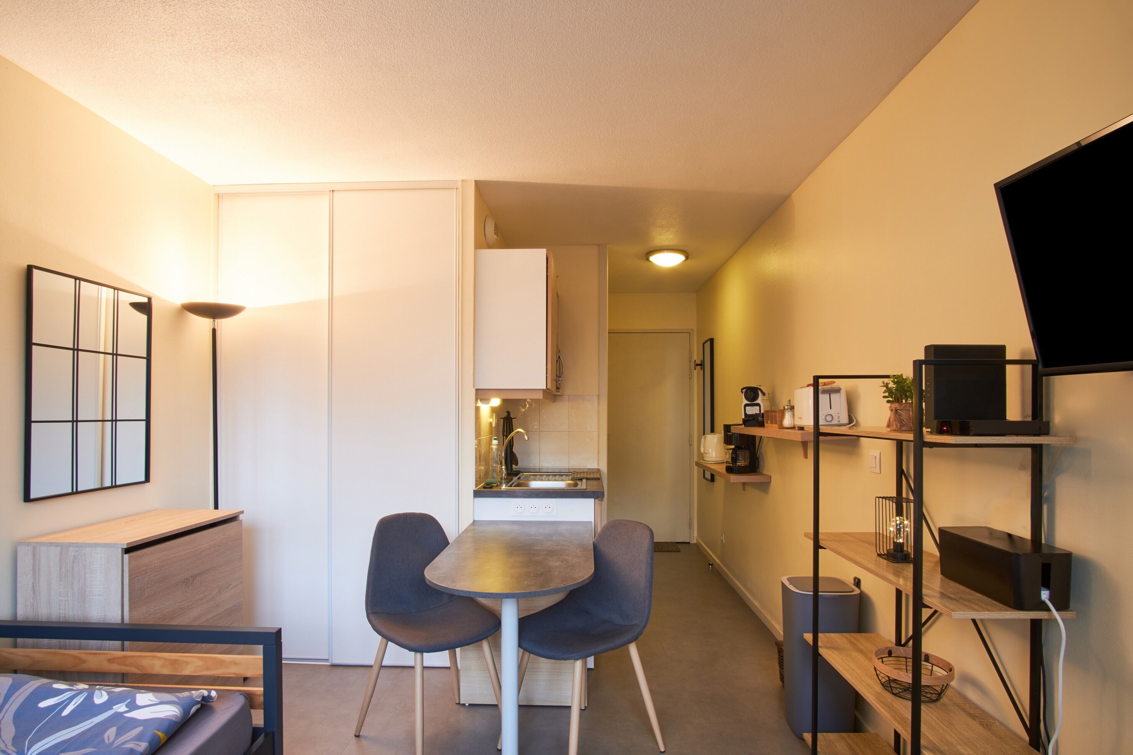 Un espace de vie compact comprenant un coin cuisine, un coin repas avec deux chaises, un lit avec une tête de lit et un téléviseur fixé au mur.
