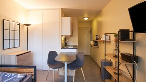 A compact living space with a kitchenette, a dining area with two chairs, a bed with a headboard, and a TV mounted on the wall.