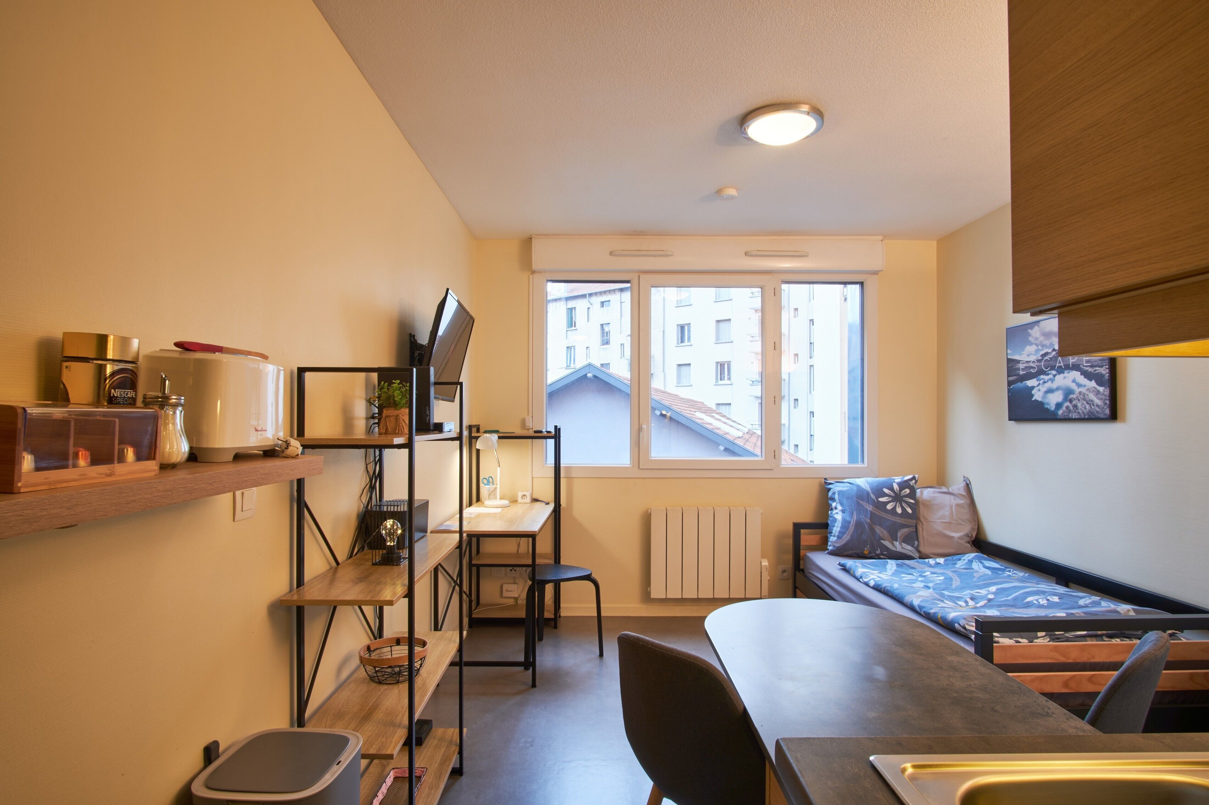 Une chambre compacte comprenant un coin cuisine, un lit simple, un bureau et une fenêtre donnant sur des immeubles.