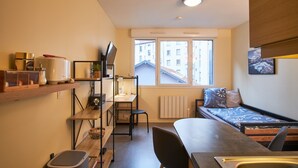 A compact room with a kitchenette, a single bed, a desk, and a window with a view of buildings.