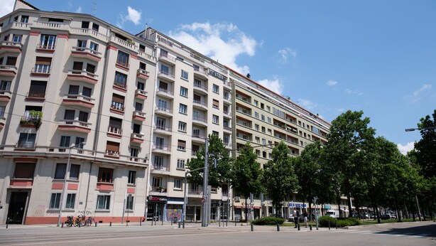 A multi-story building with balconies, a clear sky, and trees along the street.