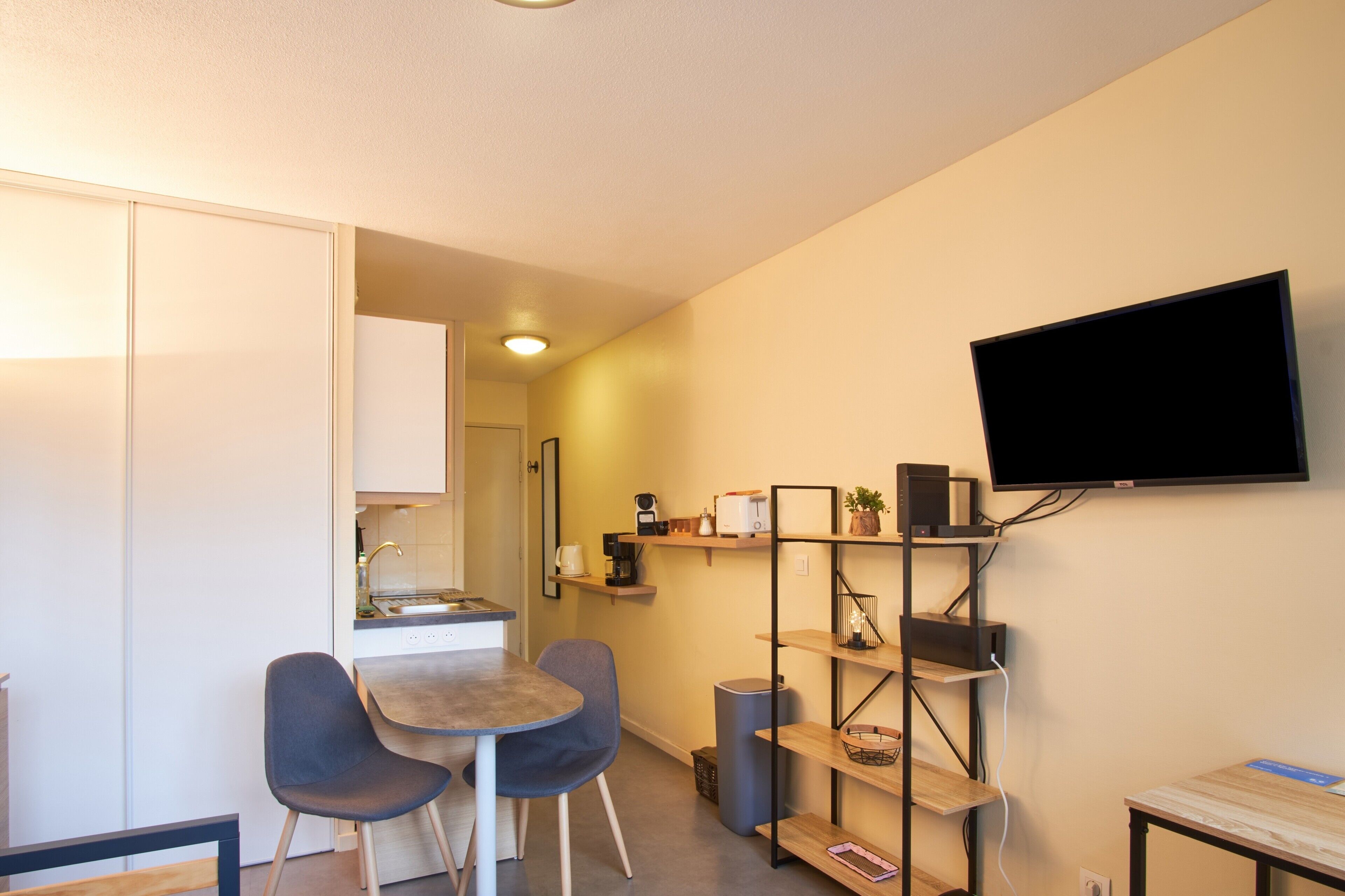 Un espace de vie compact comprenant un coin cuisine, un coin repas avec deux chaises, un téléviseur à écran plat fixé au mur et une petite étagère.