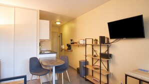 A compact living space with a kitchenette, a dining area with two chairs, a flat-screen TV mounted on the wall, and a small shelving unit.