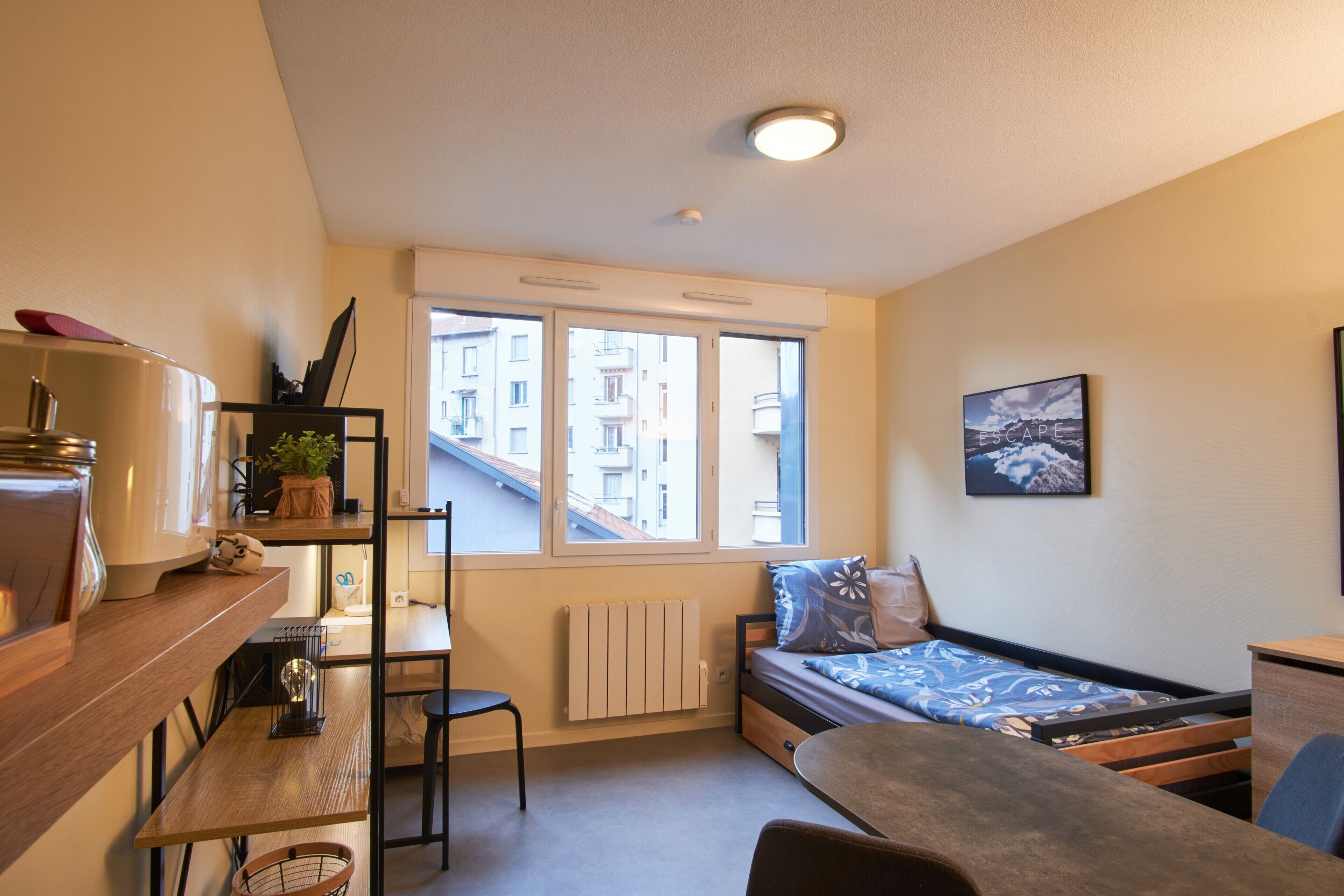 Une petite pièce comprenant un lit, un bureau, une chaise, un radiateur et une fenêtre donnant sur des immeubles.