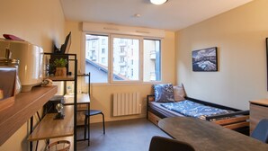A compact room with a bed, a desk, a chair, a radiator, and a window with a view of buildings.