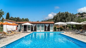 A swimming pool with lounge chairs and umbrellas, a building with large windows, and a hillside in the background. - Loft Apartment 'Marketos Studio  2' with Mountain View, Wi-Fi and Air Conditioning (Kefalonia)