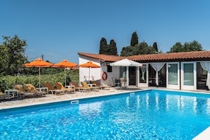 A swimming pool with sun loungers and umbrellas, a covered seating area, and a building with a tiled roof.