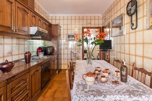A kitchen with wooden cabinets, a tiled backsplash, a dining table with a lace tablecloth, and a clock on the wall.