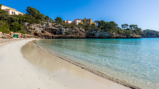 A beach with clear water, a sandy shore, and buildings on a rocky cliff. - Apartment 'Apartamento Bon Aire N1' with Shared Pool, Wi-Fi and Air Conditioning (Cala Ferrera)