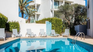 A swimming pool with lounge chairs, a tiled wall, and a building with balconies and greenery. - Apartment 'Aquamarina 7' with Shared Pool, Wi-Fi and Air Conditioning (Cala Ferrera)