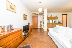 Un salon comprenant un meuble TV en bois, un canapé blanc, une bibliothèque et un coin cuisine avec des placards en bois.