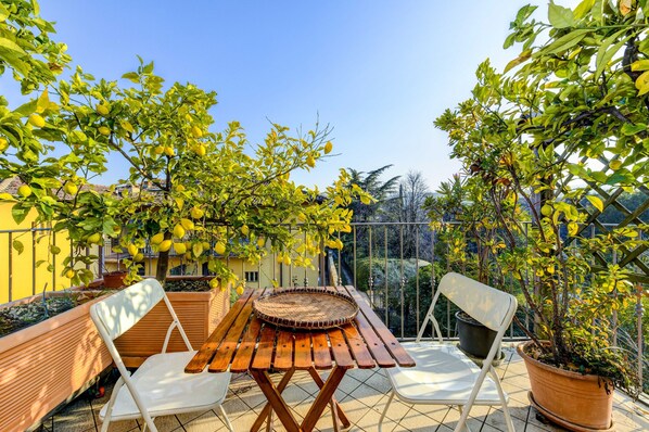 Une terrasse avec une table et des chaises en bois, entourée de plantes en pot et d’un bâtiment jaune.