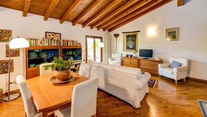 A cozy living room with a wooden ceiling, a white sofa, a dining table with chairs, a bookshelf, and a television. - Apartment 'Attic In The Center Of Salò' with Balcony, Wi-Fi and Air Conditioning (Salò)
