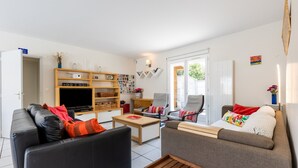 Een moderne woonkamer met een bank, fauteuil en salontafel. Er is een televisie, een boekenkast en een deur die naar buiten leidt.