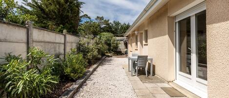Un chemin menant à un bâtiment doté d’une terrasse, d’une table avec des chaises et d’une fenêtre offrant une vue sur les arbres.