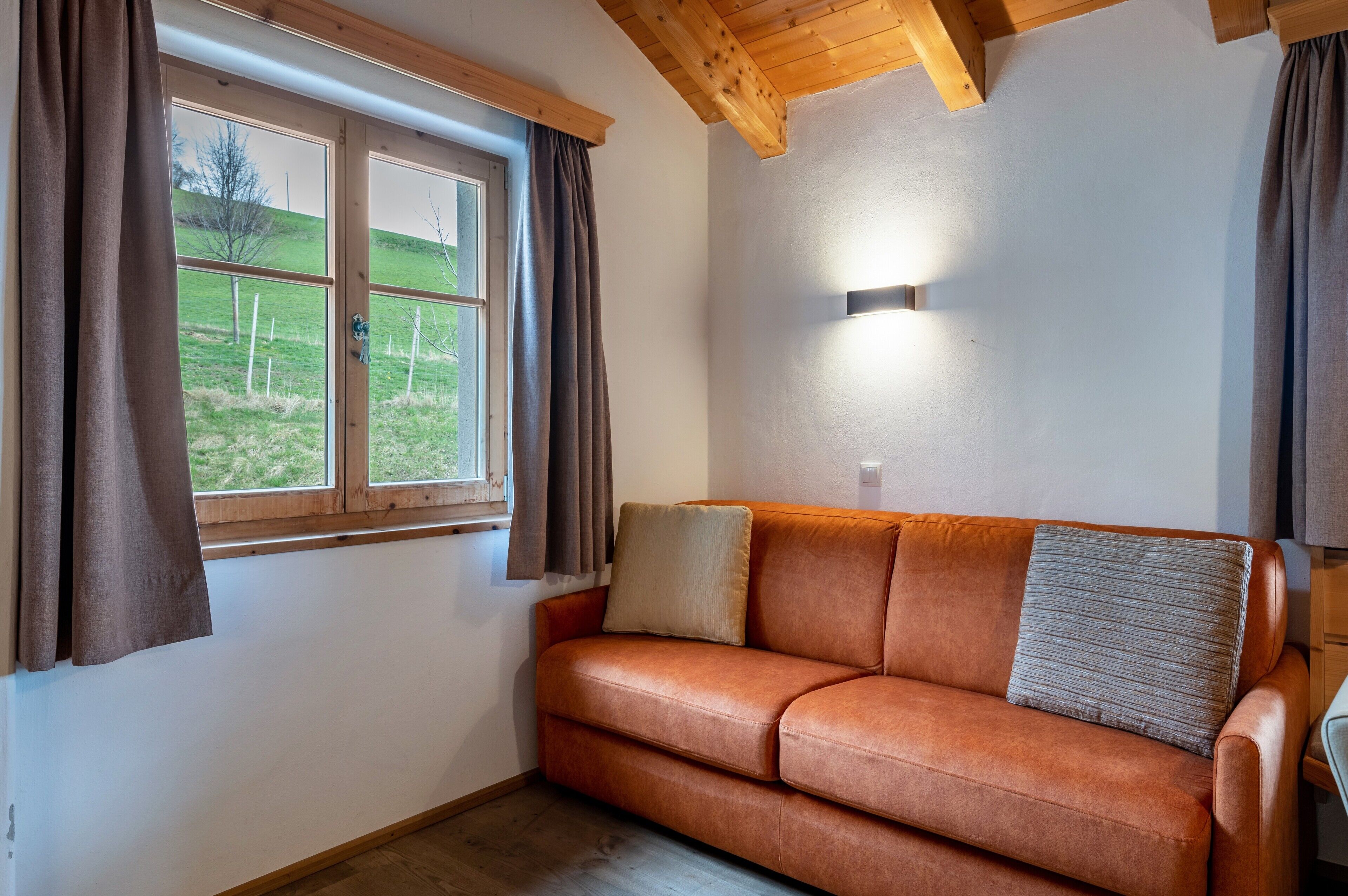 A room with a wooden ceiling, a window with curtains, a leather sofa with cushions, and a wall-mounted light fixture.