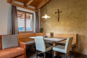 Un coin repas avec des bancs en bois et une table, une croix accrochée au mur et une fenêtre avec des rideaux.