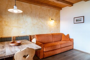 Een houten eettafel met een kom, een houten stoel met een hartvormig uitsparing, een oranje bank, een wandlamp en een lijst met een afbeelding aan de muur.
