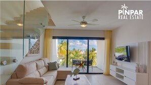 A modern living room with a sofa, coffee table, TV, and a balcony with a view of palm trees.