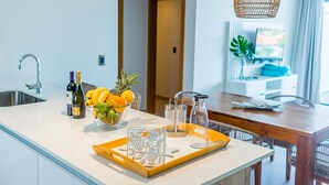 A modern kitchen with a white countertop, a stainless steel sink, a bowl of fruit, and a tray with glasses and condiments.