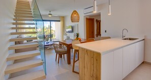 A modern kitchen with a central island, a dining area with a glass partition, and a staircase leading to an upper level.