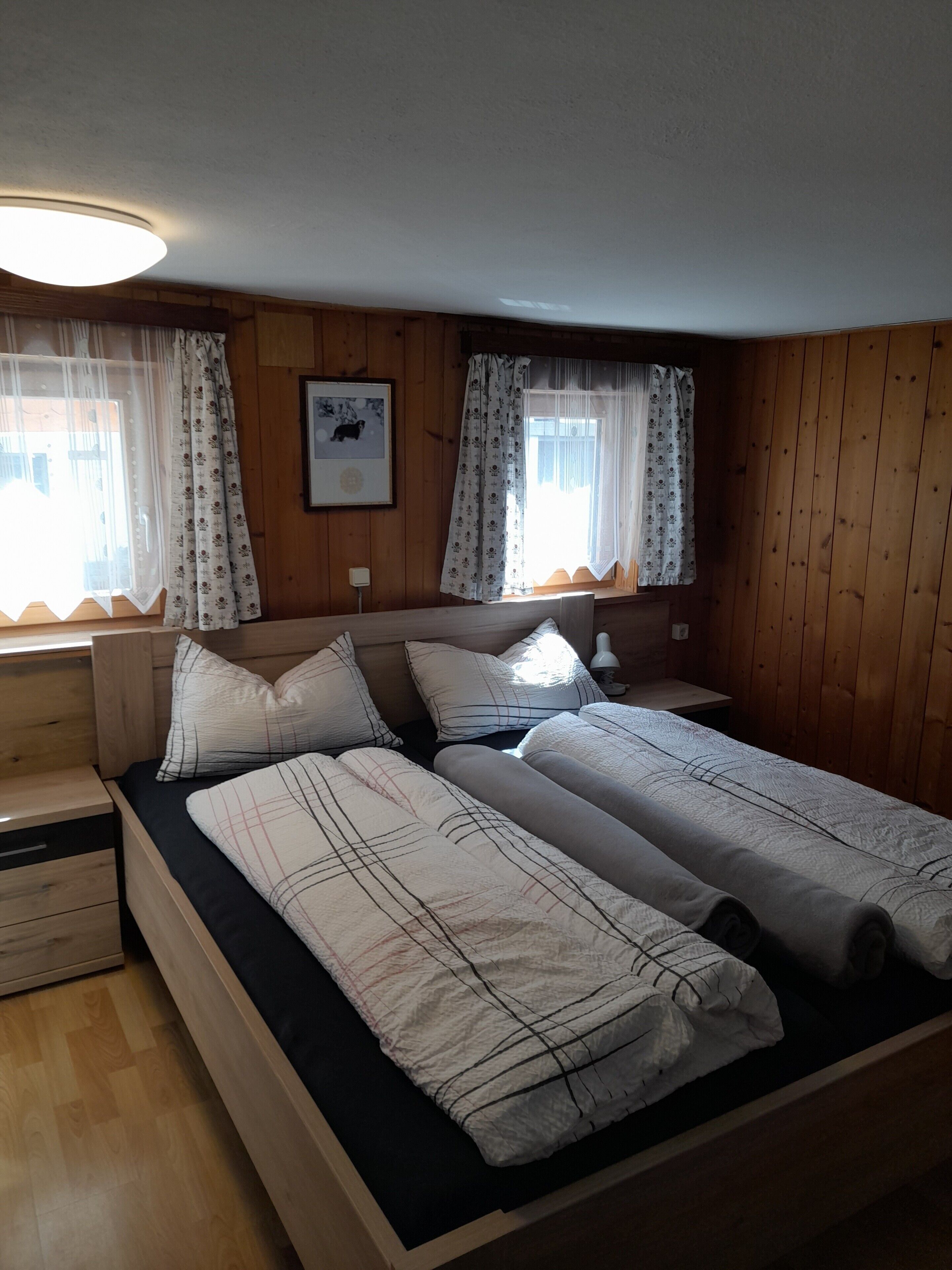 A room with two beds, wooden walls, a nightstand, a bedside table, and a framed picture on the wall.