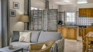 A living room with a sofa, a wicker chair, a table, and a kitchen area with wooden cabinets and a window.