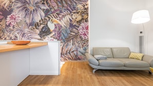 A modern living room with a wooden floor, a beige sofa, a wall with a tropical floral mural, and a white lamp.
