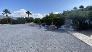 A gravel driveway leading to a building with a stone wall, surrounded by palm trees and various shrubs.
