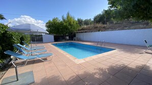A rectangular swimming pool with a tiled deck, surrounded by blue lounge chairs and a striped umbrella.