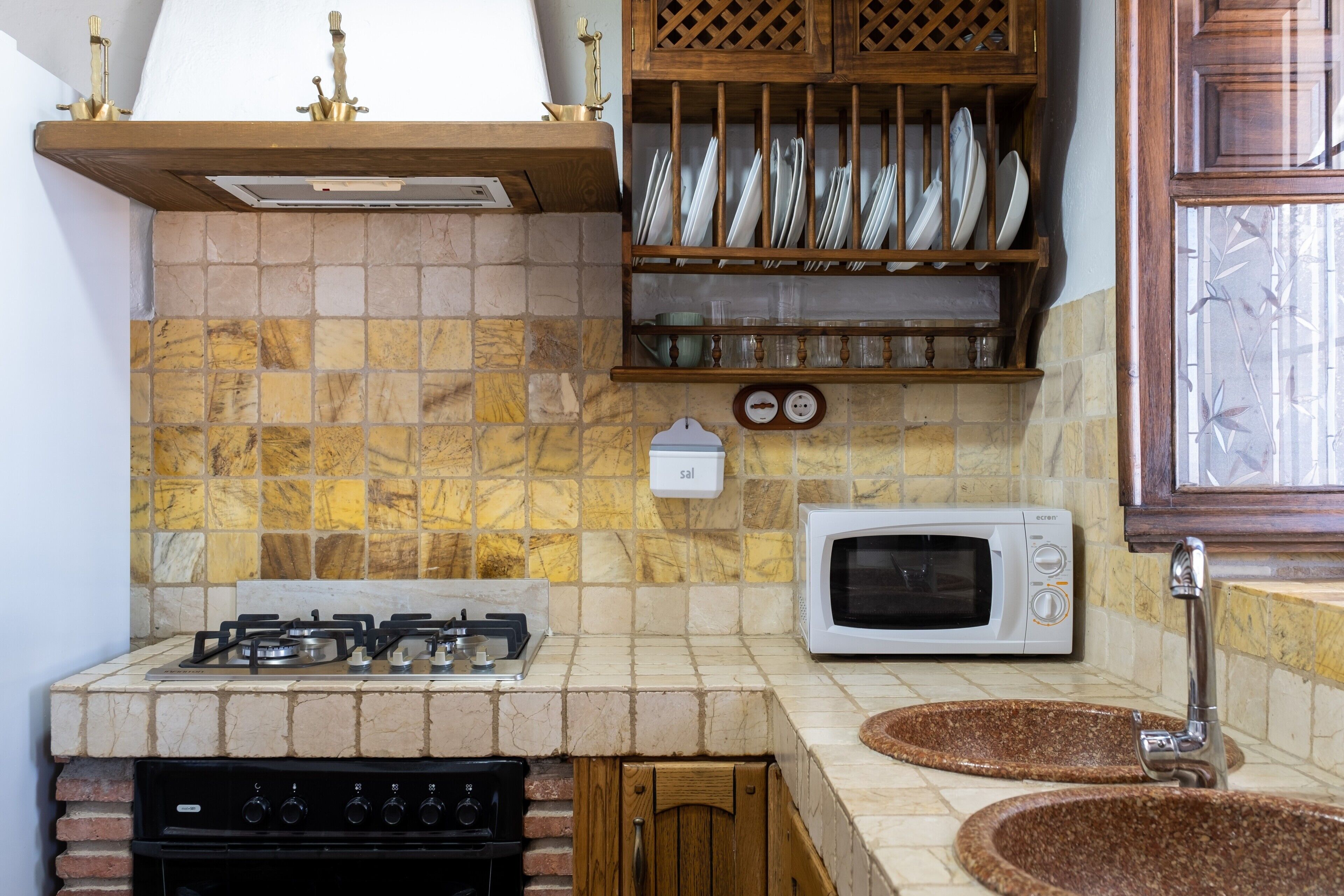 Une cuisine équipée d’une cuisinière à gaz, d’un four à micro-ondes, d’un évier et d’une étagère où sont rangés la vaisselle.