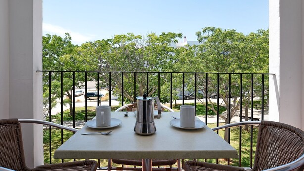 A balcony with a table set for two, a metal coffee pot, and two white cups on saucers.