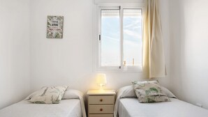 Ein kleines, helles Schlafzimmer mit zwei Betten, einem Nachttisch mit Lampe, einem Fenster mit Vorhängen und einem Wandbild.