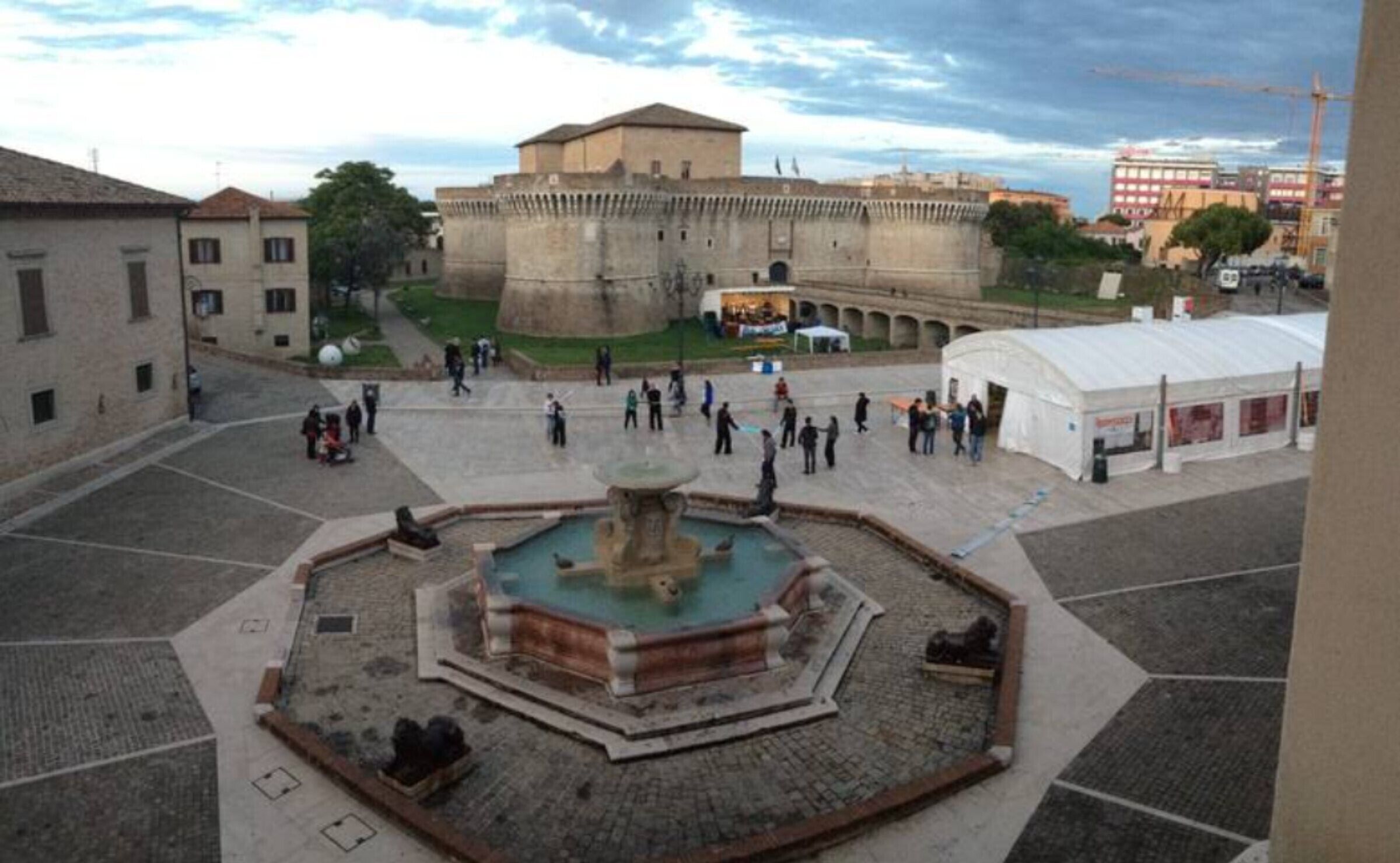 A courtyard with a fountain, a large building in the background, and a white tent-like structure.