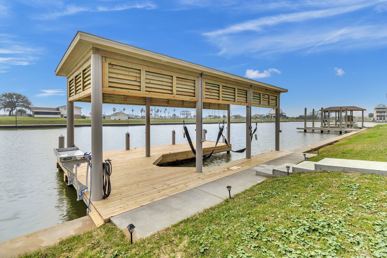 A wooden dock with a covered seating area extending over a body of water, supported by concrete pillars.