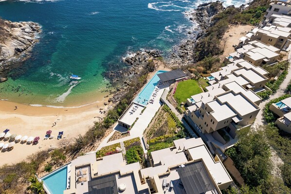 An aerial view of a coastal hotel with a swimming pool, beach, and rocky shoreline. - Penthouse | Views + Beach Access + Rooftop Terrace (Huatulco)