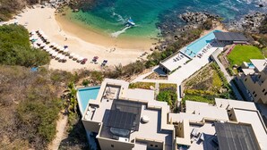 An aerial view of a coastal resort with a sandy beach, a swimming pool, and multiple buildings. - Penthouse | Views + Beach Access + Rooftop Terrace (Huatulco)
