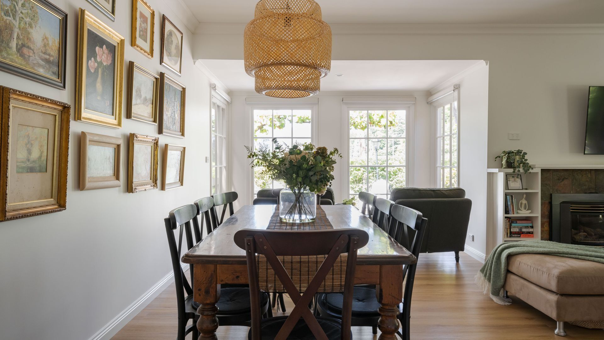A dining area with a wooden table, chairs, and a vase of flowers. There are framed pictures on the wall and a television on the right.