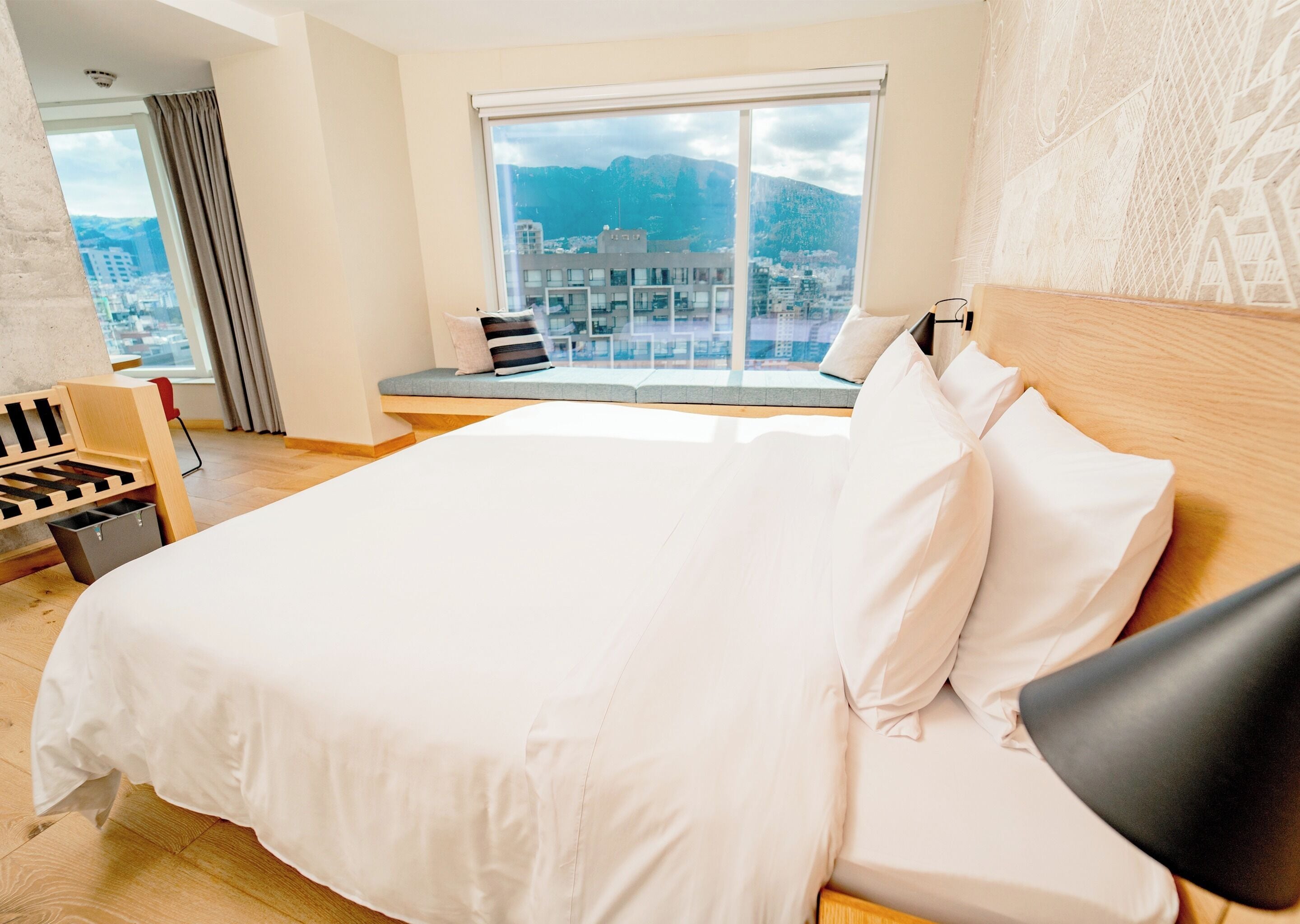 Quarto de hotel com uma cama grande, vista para a cidade e um espaço de estar com um banco e cadeiras.